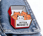 Enamel pin with cartoon cats in a box labeled 'Kitten Nuggets' on denim fabric © Tigoli® 2026 All Rights Reserved Panda Loco Ltd 